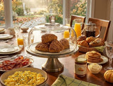 A beautifully set dining table in a cozy autumn kitchen, featuring a breakfast spread with croissants under a glass dome, scrambled eggs, bacon, pancakes, orange juice, and small pumpkins, with a large window showing an outdoor fall garden view.